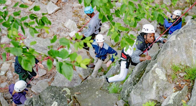 Looking down, you can see several people wearing helmets standing at the base of a rock wall. One person is climbing. 